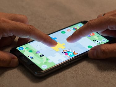 Close-up of fingers tapping a casual puzzle game on an Android phone screen, ambient indoor light, phone resting on a fabric surface, no glamour lighting