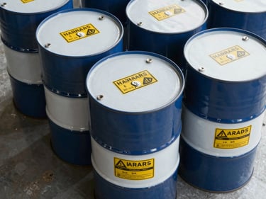 Close-up overhead shot of stacked industrial chemical drums with standardised hazard labels on a warehouse floor, daylight from above, clean steel surface, sharp detail on container markings