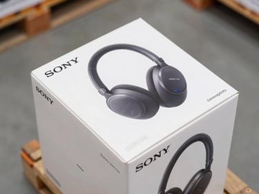 Close-up overhead view of sealed Sony WH-1000XM and Samsung Galaxy Buds retail boxes stacked on a pallet shelf, serial number stickers visible on box edges, clean warehouse daylight from above, plain concrete floor in background