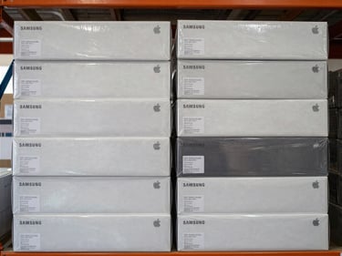 Overhead close-up of neatly stacked sealed Samsung and Apple case boxes arranged in two rows on a warehouse shelf, printed authentication seals visible, clean daylight from the left, neutral background