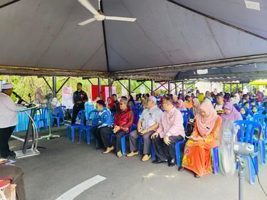 Community members gather under a tent for an outdoor speech at a local Malaysian event.