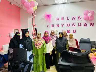 A group of women in hijabs attend a breastfeeding class with pink balloons and wall decor.