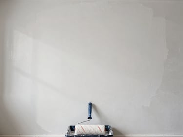 Overhead shot of a freshly painted white drywall section, paint roller resting on a tray at the bottom edge, natural daylight from a high window casting soft shadows