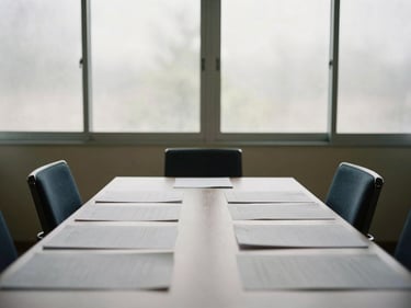 Empty quiet meeting room with two chairs facing each other across a narrow table, documents spread flat, natural daylight through frosted glass partition, no people visible
