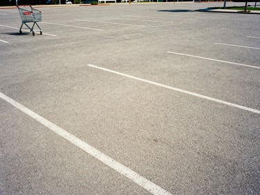 Empty parking lot at midday in hard summer sun, early 2000s, digital grain, a lone shopping cart at the far edge, flat harsh shadows