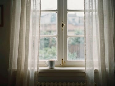 An early 2000s digital photograph of a living room window, morning light coming through thin curtains, a coffee mug on the sill, slightly flat digital color rendering, no grain