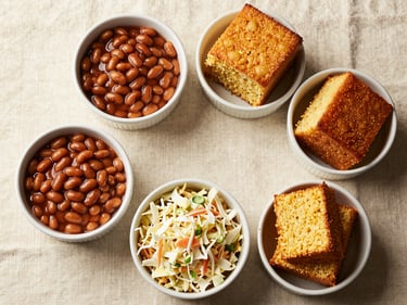 Overhead flat-lay of classic BBQ side dishes in white ceramic bowls on a cream linen surface — baked beans, coleslaw, cornbread — warm natural window light