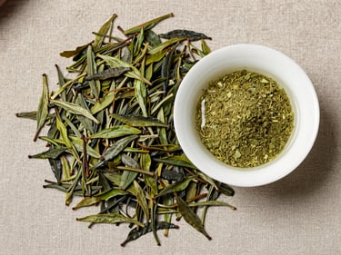 Flat-lay of loose green tea leaves and a small dish of EGCG extract powder on warm gray linen, diffused natural daylight from above, tight overhead crop, no bottles or labels
