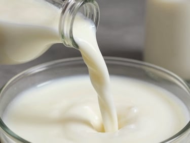 Intimate close-up of fresh whole milk being poured from a steel ladle into a clear glass bottle, natural daylight catching the white stream mid-pour, cream-coloured surface beneath, no background clutter