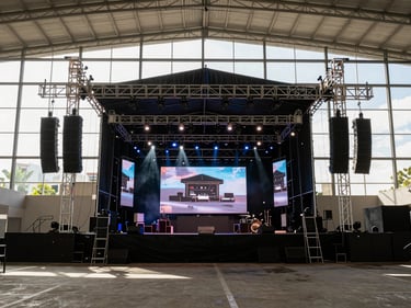 Wide environmental shot from the back of house looking toward a fully-rigged concert stage in a large Monterrey venue — speaker arrays flanking both sides, moving-head lights pointing at the stage, LED screen panels displaying live content at center, the room ready before doors open, crisp daylight from venue windows cutting across the rigging, no performers visible