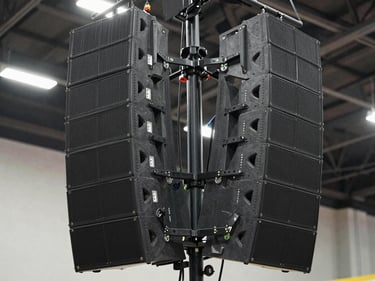 Close-up to mid-shot of a professional line-array speaker cluster mounted on a truss above a Monterrey venue stage, shot from slightly below looking up — multiple speaker cabinets stacked symmetrically, rigging hardware visible and in focus, venue ceiling structure above, even studio-bright lighting revealing the precision of the installation, no people in frame