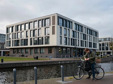 Environmental shot of a Dutch university campus — a modern low-rise faculty building beside a canal, a student at a bicycle rack removing a backpack, flat grey sky, cool diffuse daylight, calm urban academic context