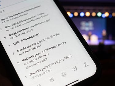 Overhead phone shot showing a Q&A submission interface with Vietnamese text questions listed, event stage blurred in background, ambient stage lighting