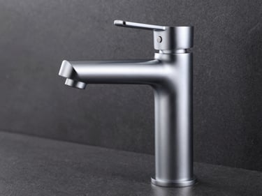 Close-up studio shot of a freestanding chrome bathroom faucet on a dark anthracite surface, sharp modeling light from upper left revealing the cylindrical neck and lever handle finish, tight product framing, no background detail