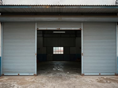 Warehouse entrance with wide roller shutter open, concrete floor visible inside, Auto Nagar district, overcast daylight, no people, medium shot showing facade and forecourt