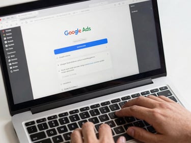 Close-up overhead of a laptop screen displaying a Google Ads campaign interface mid-edit, a hand resting near the keyboard, clean studio lighting with no glare, screen content fully readable