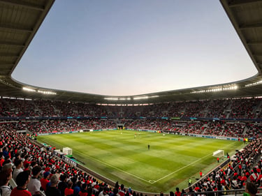 Wide shot of a packed soccer stadium at dusk, floodlights on, crowd in team colors filling the stands, golden light on the pitch