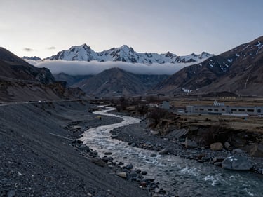 Wide environmental shot of Spiti Valley at early morning, high-altitude river cutting through grey scree, snow-dusted peaks in the far distance, low horizontal cloud layer, no people