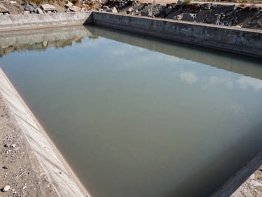 Mining site water treatment settling pond, grey sediment-laden water in a wide concrete basin, overhead medium shot showing water clarity gradient from inlet to outlet under open sky