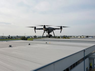Drone hovering above large industrial warehouse roof, operator visible below in PPE, overcast sky, wide environmental shot showing full roof scale