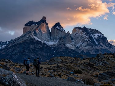 Wide shot of Torres del Paine granite spires at dusk, two hikers with backpacks paused on a ridge in the lower left, cold blue light and streaks of orange cloud above, Patagonian steppe visible below