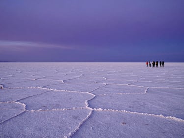 Wide environmental shot of the Atacama salt flats at blue hour, the horizon perfectly flat and infinite, a small cluster of travelers standing at the edge of the flats to the right, deep violet sky above