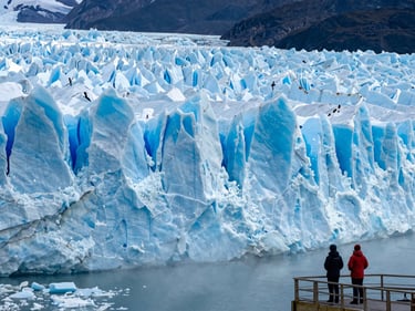 Close wide-angle of the Perito Moreno glacier face at early morning, blue ice wall filling the frame, two travelers in cold-weather gear standing at the viewing platform lower right, mist rising off the water