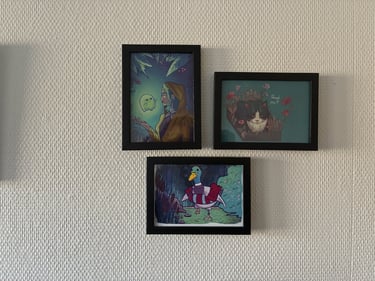 Three framed illustrations of a ghost girl, a cat in flowers, and a duck on a white wall.