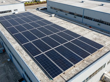 Overhead drone shot of a rooftop solar array on an industrial warehouse, panels arranged in tight rows across a flat concrete roof, harsh midday light casting minimal shadows, neutral urban context