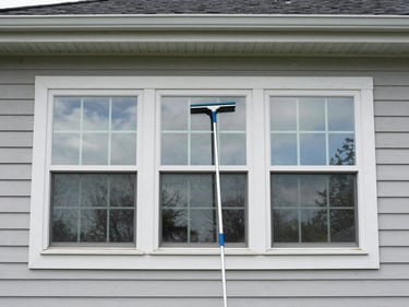 Wide shot of a residential two-story window being cleaned by a professional with a squeegee, clear reflection of sky visible on the cleaned pane, bright even daylight, Maryland suburban home exterior, documentary framing