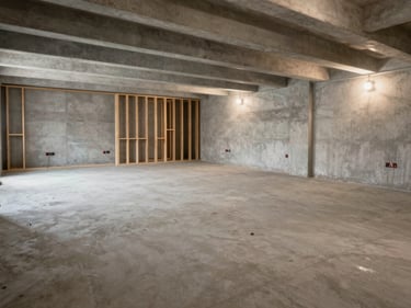 Wide shot of a basement mid-renovation, concrete floor newly poured, framing studs visible against raw concrete walls, work lights casting even natural-tone illumination