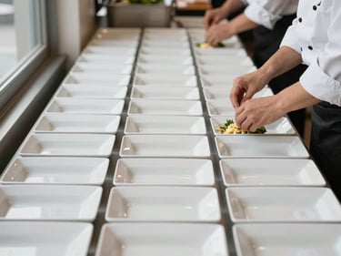 Overhead shot of a corporate buffet service line: identical white rectangular dishes arranged in precise rows on a long table, hands in chef whites adjusting a garnish, soft diffused daylight from a window left of frame