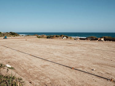 Ground-level view of a marked building site adjacent to Asila coastline, Atlantic sea visible in background, surveying stakes visible, natural midday light, no people