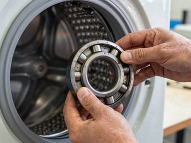 Extreme close-up of a technician's hands lifting the drum bearing from a disassembled washing machine on a workshop bench, natural side-light, grease and metal visible, no faces, tight frame on the mechanical components