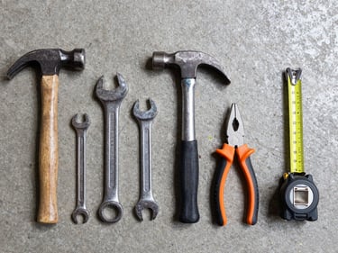 Head-on flat lay of hand tools — hammers, wrenches, pliers, and tape measures arranged in a row on a concrete floor, bright even warehouse lighting, no shadows obscuring tool markings