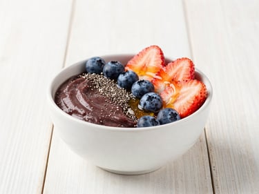 A beautifully styled acai bowl topped with sliced strawberries, blueberries, chia seeds, and a drizzle of honey, natural wood table background