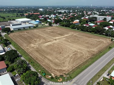 Wide aerial drone shot of undeveloped land parcel adjacent to a main road in Johor Bahru, visible lot perimeter, cleared ground, industrial surroundings, overcast natural daylight, no people