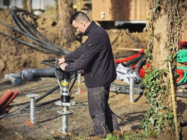 A construction worker installing a screw pile foundation for a new building using a handheld hydraulic drive.