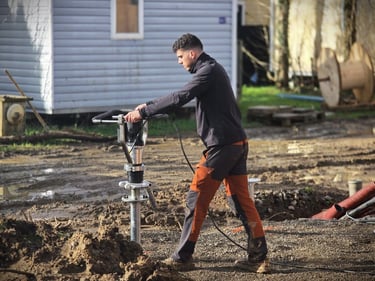 A construction worker using a power driver to install helical screw piles into muddy ground.