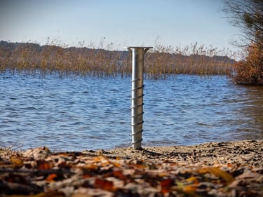 Galvanized steel ground screw anchor installed in sandy shore for lakeside dock foundation.