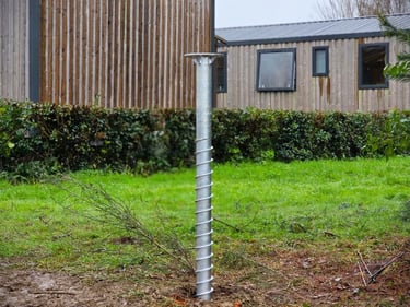 Galvanized steel ground screw foundation installed in a grassy backyard near wooden garden buildings.