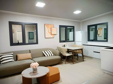 Modern studio apartment with a grey sofa, minimalist dining set, and white kitchenette with a refrigerator.