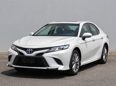 Three-quarter front view of a clean 2019 Toyota Camry in white on a bright outdoor lot under even daylight, neutral asphalt background, no people