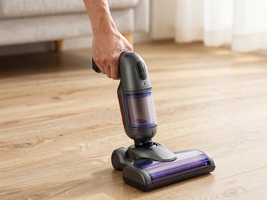 Cordless handheld vacuum cleaner held in a person's hand over a wooden floor, bright natural indoor light, device in sharp focus