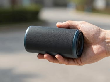Person holding a compact wireless Bluetooth speaker in one hand outdoors on a sunny day, product details sharp and color-accurate