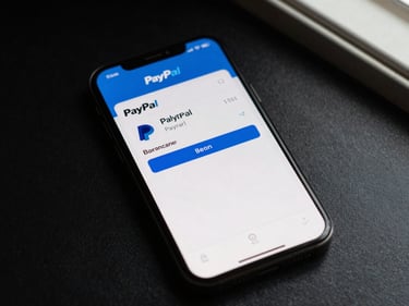 Overhead flat-lay of a smartphone screen showing a PayPal balance summary, natural soft window light, dark surface beneath, no hands, documentary close-up