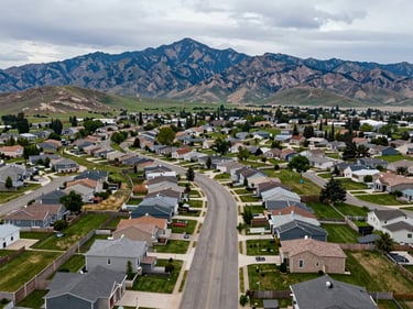Aerial view of Riverton Utah neighborhood, curving residential streets, Wasatch peaks in background, overcast daylight, green parks and modern homes visible