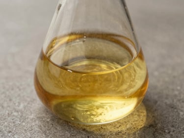 Extreme close-up of castor oil in a clear glass laboratory flask, north-facing daylight, warm amber refraction, stone surface, no people