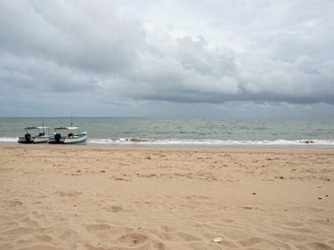 Panoramic overcast-light shot of Nagaon beach, wide flat sand in the foreground, grey-green sea meeting a low cloudy horizon, a pair of fishing catamarans pulled ashore on the left edge, no tourists visible