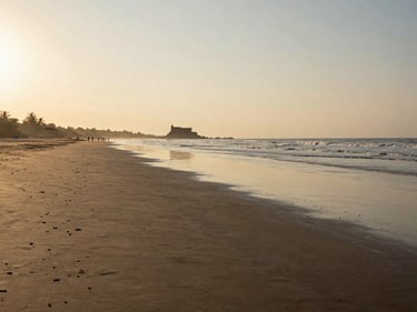 Wide environmental shot of Alibaug beach at golden hour, the shoreline stretching toward Kolaba Fort silhouette in the shallow water, warm low light raking across wet sand, sparse early-morning walkers in the far distance, no posed subjects